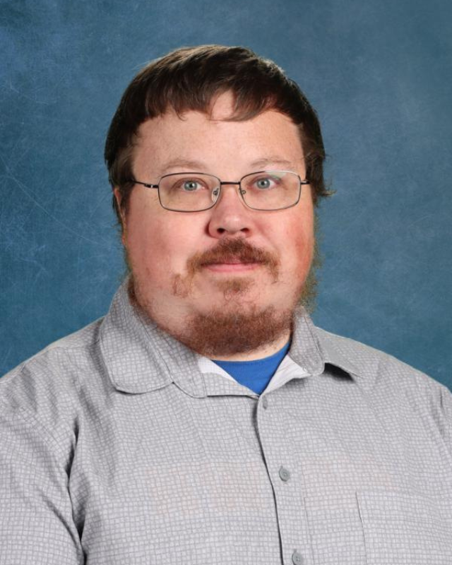 Portrait of a man with short dark hair, glasses, and a goatee/beard wearing a light gray checkered shirt against a blue backdrop.