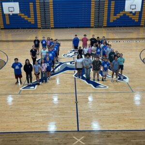 Group of students standing on a gym floor around a large Mustang mascot logo, with blue and gold bleachers in the background.