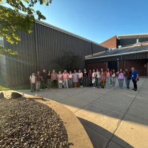 A line of people standing outside a brick and metal-paneled building on a sunny day.