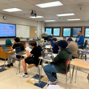 Classroom scene with students at desks using laptops, while a teacher stands at the back near windows and a projector screen is displayed on the wall