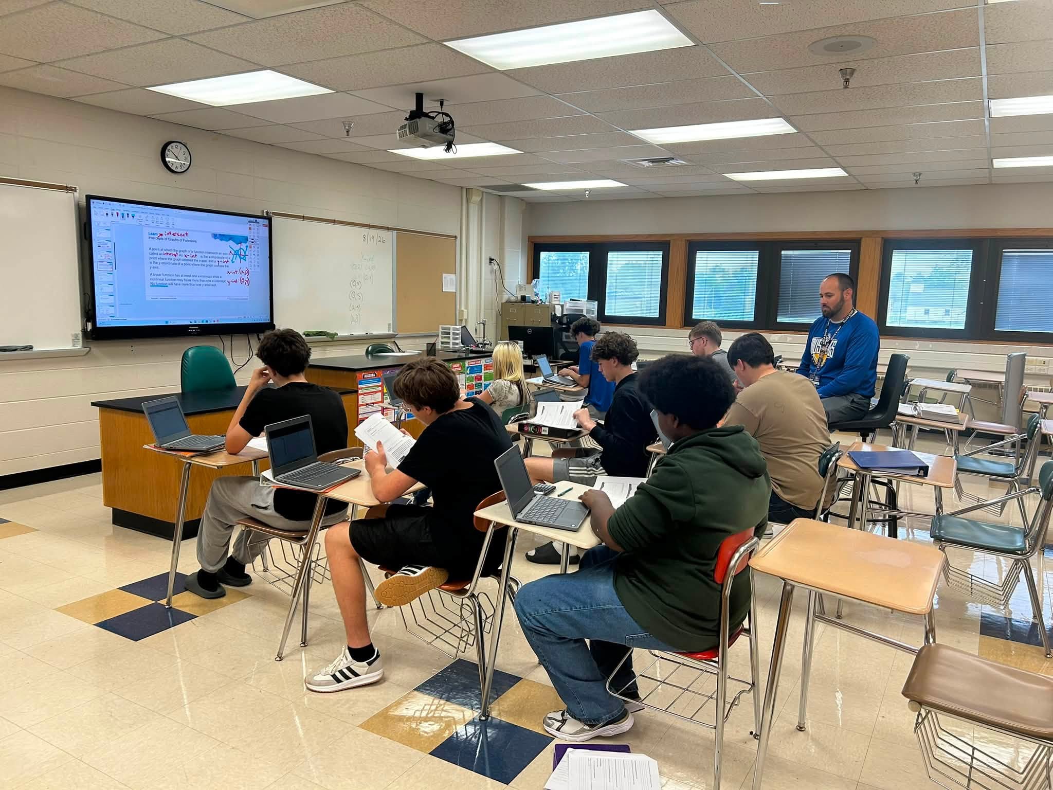 Students at desks using laptops in a classroom, with a teacher standing near the back by windows.