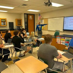 Teacher in a blue shirt stands at the whiteboard, teaching a math lesson to students with laptops in a classroom.",