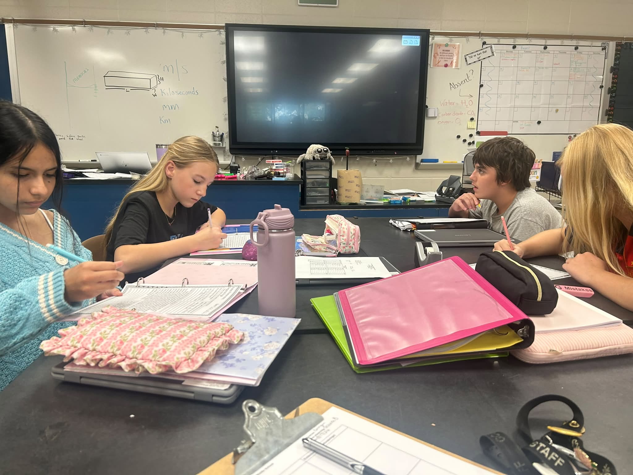Students seated around a classroom table, writing in notebooks with a large screen on the back wall.
