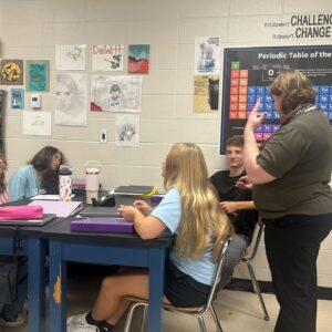 Teacher standing and gesturing over a group of students seated at a lab table in a classroom, with a large Periodic Table poster on the wall