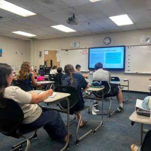 Classroom with students seated at desks, facing a projected slide on the wall screen.