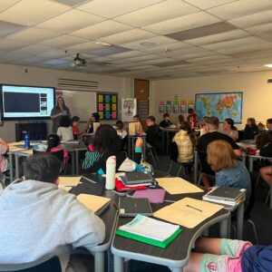 Teacher presenting at the front of a classroom as students sit at tables with notebooks and laptops, a projector screen visible.