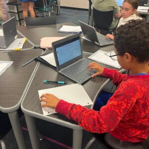 Student in a red patterned hoodie sits at a desk typing on a Chromebook while a notebook lies open in front of him in a classroom. Other students at laptops are visible in the background near a science station and filing shelves.