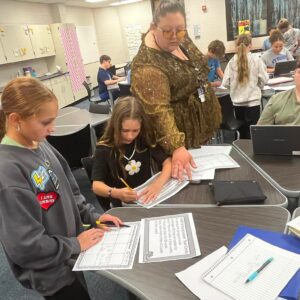 A classroom scene with students at tables, a teacher in a shimmering gold top assisting a student with a worksheet in the foreground.