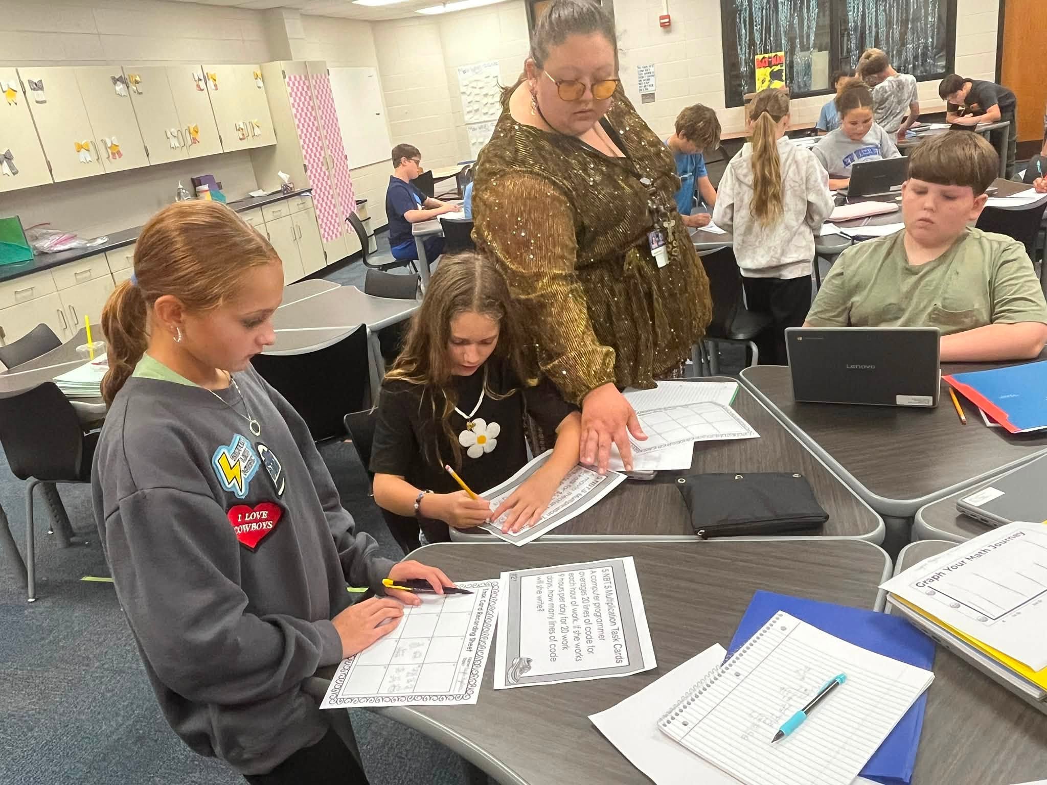 Students sit at tables in a classroom, working on worksheets while a teacher assists a student nearby.