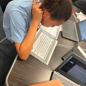 Student in a light blue shirt leans over a desk, writing on a worksheet beside an open laptop.