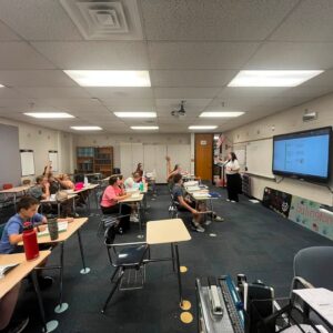 Classroom scene with students at desks, some raising hands, and a teacher at the front near a large display screen.