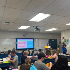 Students sit at lab tables doing worksheets while a teacher presents from the front near a blue display screen in a classroom with an American flag visible on the left.