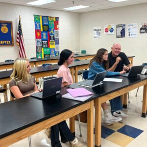 Group of students sit at long black-topped lab tables with laptops in a classroom, banner reading 'FFA Washington Chapter' and an American flag on the wall behind them.