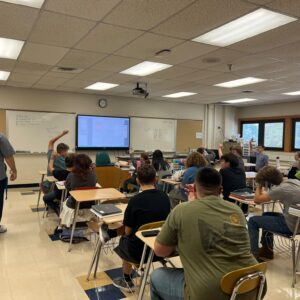 Teacher at front of classroom as students raise hands; projector screen on whiteboard and a wall clock visible.