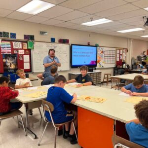 Teacher leading a class at the front of an art room with students at tables and a whiteboard behind him.