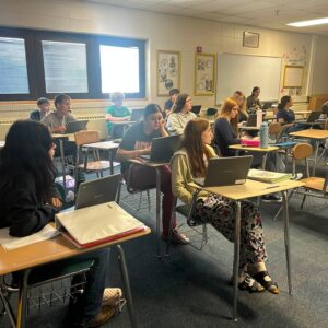 Students sit at individual desks in a classroom, each with a laptop open, listening to a lesson.