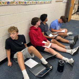 Four students sit along a school hallway wall with laptops and notebooks, writing and looking at screens during classwork.