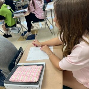Classroom scene with students at desks; a student in a bright green hoodie with cartoon eyes on the back sits at a desk while another student in a pink hoodie writes on a worksheet. A pencil case and notebooks are visible on the desks.