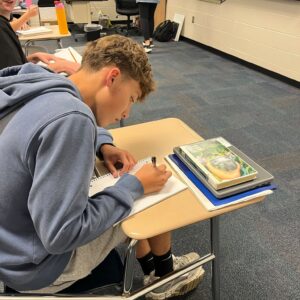 Student in a blue hoodie writes in a notebook at a desk in a classroom; other students and a teacher stand near a whiteboard in the background.