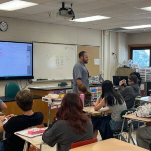 Teacher standing at the front of a math classroom as students work at desks, with a projector screen and whiteboard behind him.