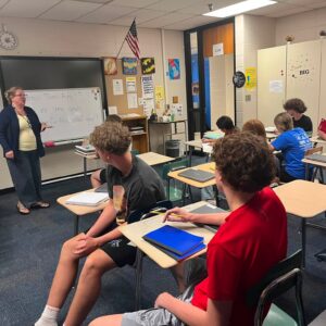 Teacher stands at the whiteboard delivering a lesson to students seated at desks in a classroom.