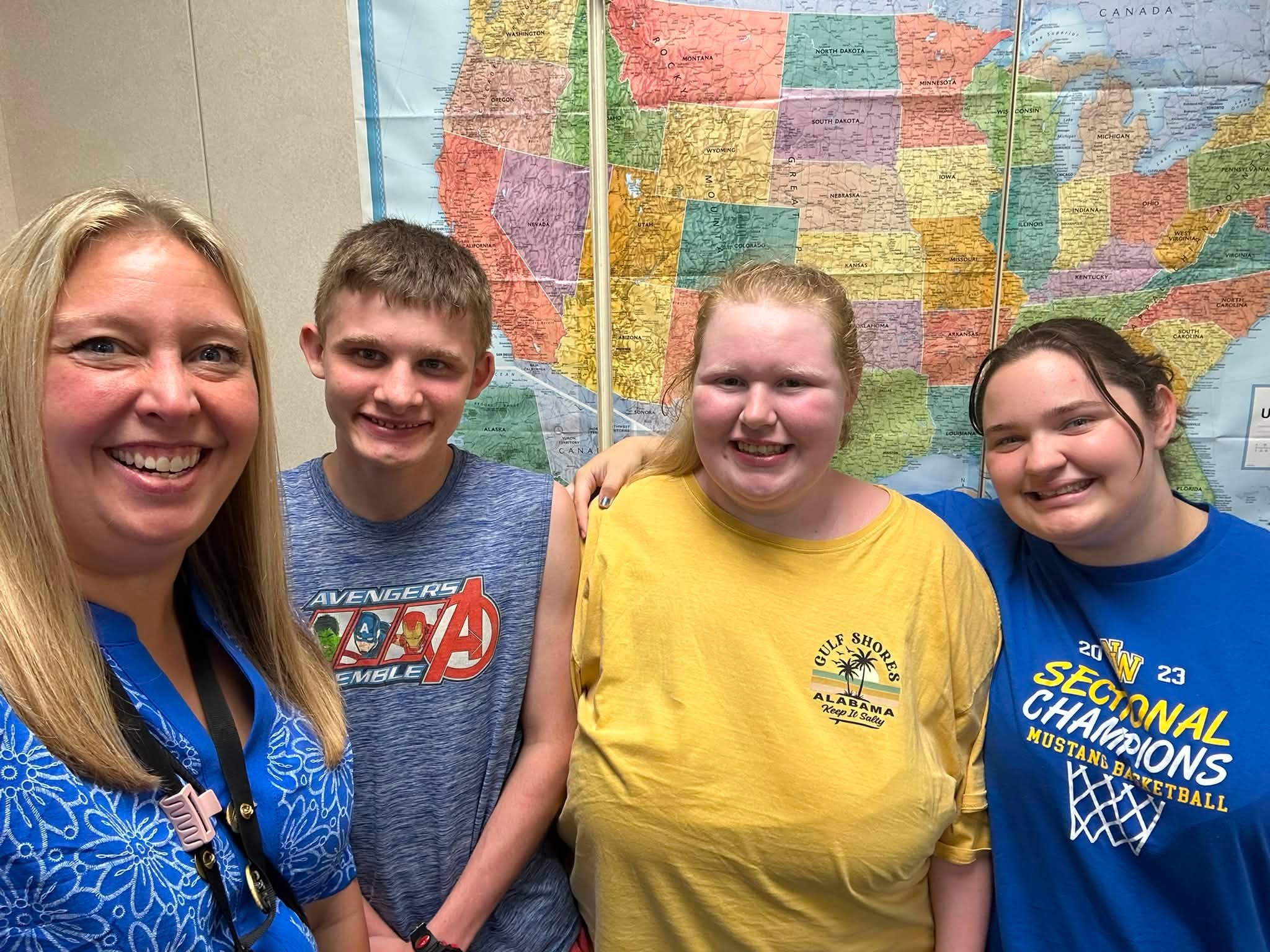 Four people smile for a group selfie in front of a large colorful United States map on the wall behind them, likely in a classroom or office.