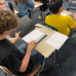 Students seated at desks in a classroom, working on laptops and writing in notebooks with papers and folders nearby.