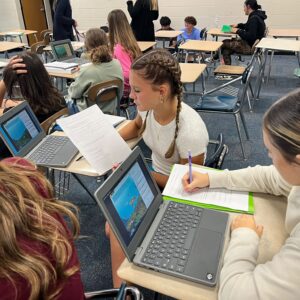Students in a classroom working on laptops at scattered desks, with papers and notebooks nearby.