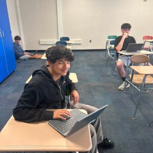 Student in the foreground smiles while typing on a laptop at a desk in a classroom filled with other students using laptops at desks in the background.