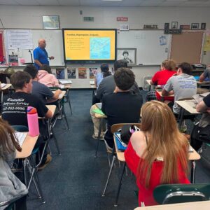 Teacher stands at front of a classroom presenting to students, with a map slide projected on a yellow screen.