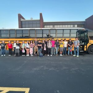 Group of students standing in a parking lot in front of a yellow school bus and a brick school building with clear blue sky above.