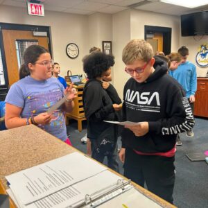 Group of students in a classroom gathered around a counter with open binders, a girl in a purple shirt speaking while a boy in a NASA hoodie reads a sheet of paper.