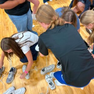 People kneeling and crouching on a wooden gym floor, tying sneakers, with many gray athletic shoes and white laces spread around; a dog on a purple leash in the background.