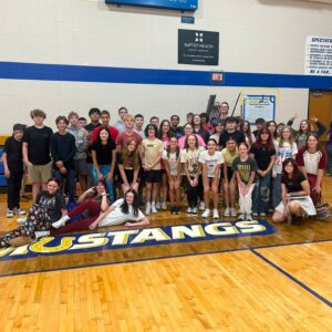 Large group of students posing for a group photo in a gymnasium, with 'MUSTANGS' on the wooden floor in front of them.