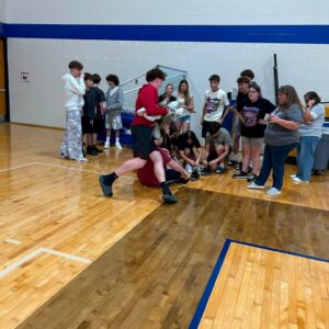 Group of teenagers watching a chaotic moment as two boys wrestle on the gym floor, others standing along the wall.