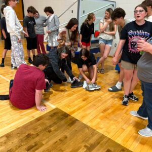 Group of teens in a gym, some kneeling to tie sneakers on a wooden floor while others stand and chat near a basketball court area.