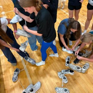 Students in a gym tying the laces of white sneakers while others watch, with many shoes scattered on the polished wooden floor.