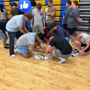 Group of students in a gym tying their athletic shoes on a wooden court, with blue and yellow bleachers in the background and a blue thumbs-up icon nearby.