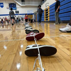 Inside a school gym, a row of sneakers tied together with laces lines the polished wooden floor toward the camera while students stand and walk near blue bleachers in the background.