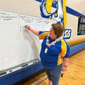 Woman in a blue and yellow polo writes on a large whiteboard in a school gym, with blue-and-yellow mural and bleachers in the background.