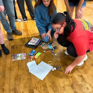Two girls kneel on a gym floor surrounded by school supplies, studying papers and calculators laid out in front of them while others stand nearby.