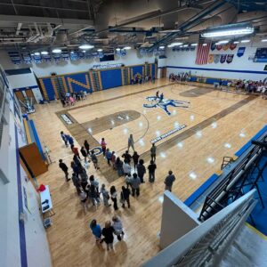 School gymnasium with a large Mustang logo on the floor; groups of students line up along the bleachers by blue and yellow seating area, banners overhead