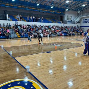 Crowd of students seated on blue and yellow bleachers watches performers dancing on a polished gym floor near orange cones in the foreground.