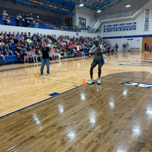 Full gymnasium with students seated on blue bleachers watching a runner navigate orange cones on a polished wood floor.
