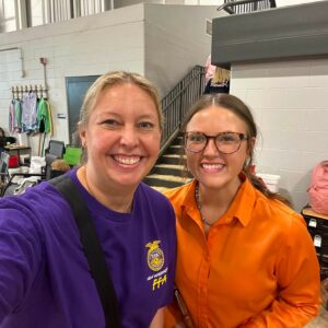 Two women smiling for a selfie in a gym-like indoor space, one in a purple FFA shirt and the other in orange, with chairs and coats in the background.