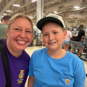 Smiling woman in a purple shirt and a boy in a blue Carhartt shirt take a selfie at a crowded indoor event with bleachers and metal rails in the background.